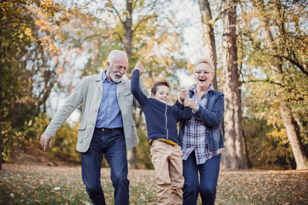 Grandparents and their grandson walking outside in a wooded park, all holding hands while smiling and laughing.